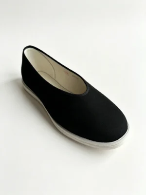 LUNO LN-M3024 traditional Chinese cloth shoes angled side view - showcasing a sleek black canvas upper with a rounded toe and elastic side panels for easy slip-on wear, paired with a white layered rubber sole; the interior reveals a quilted insole with embroidered mugwort leaves and red "Beijing Cloth Shoes" characters, emphasizing herbal comfort and cultural craftsmanship.